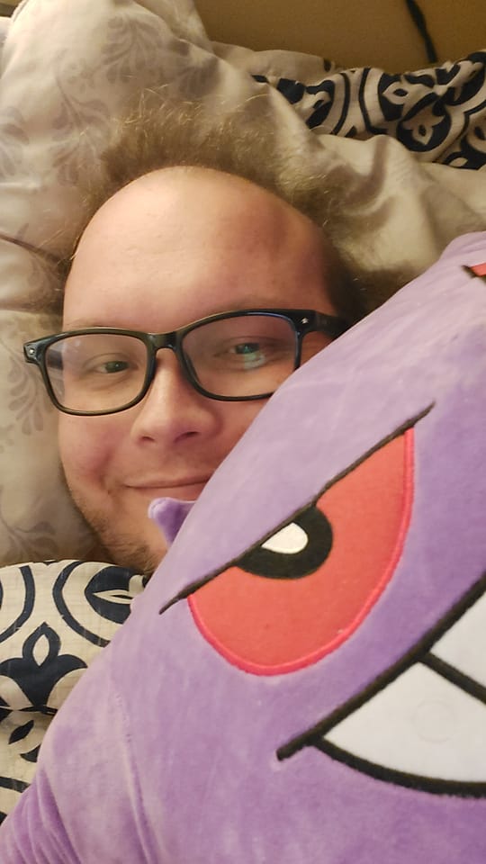 A man with thin green hair lies in bed smiling and holding a large Gengar plush 